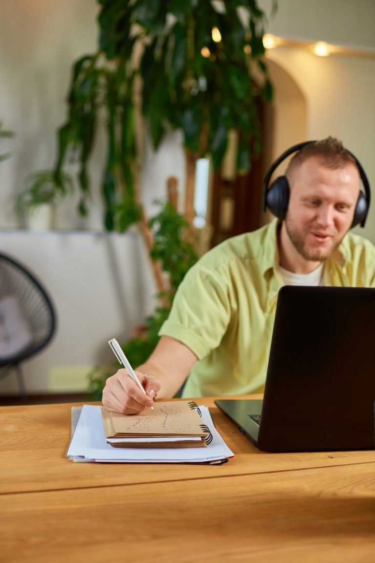 Man met koptelefoon volgt online training en maakt aantekeningen aan tafel
