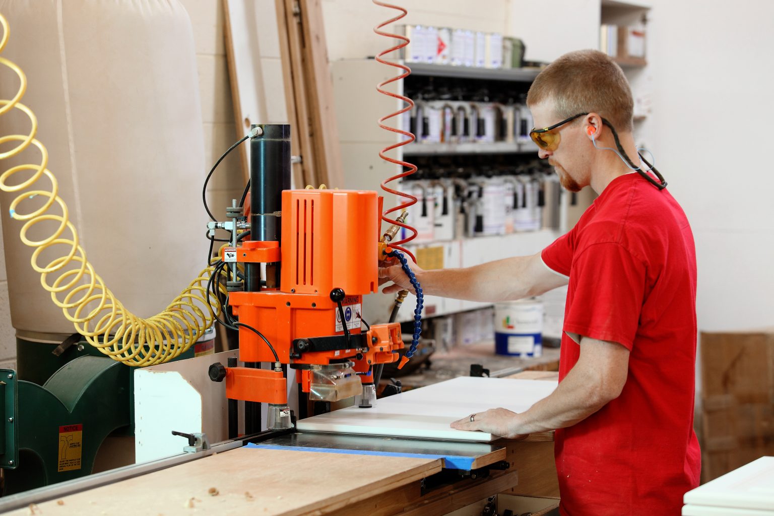 Man met veiligheidsbril en gehoorbescherming die een houtbewerkingsmachine bedient in een werkplaats.